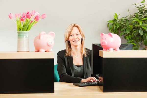 A happy, smiling Caucasian woman bank teller working with a computer with financial information tables on the screen. She sits behind the bank counter window decorated with pink piggy banks, she is looking at the camera posing in a retail bank office building. Photographed from the customer side of the counter in a horizontal format.